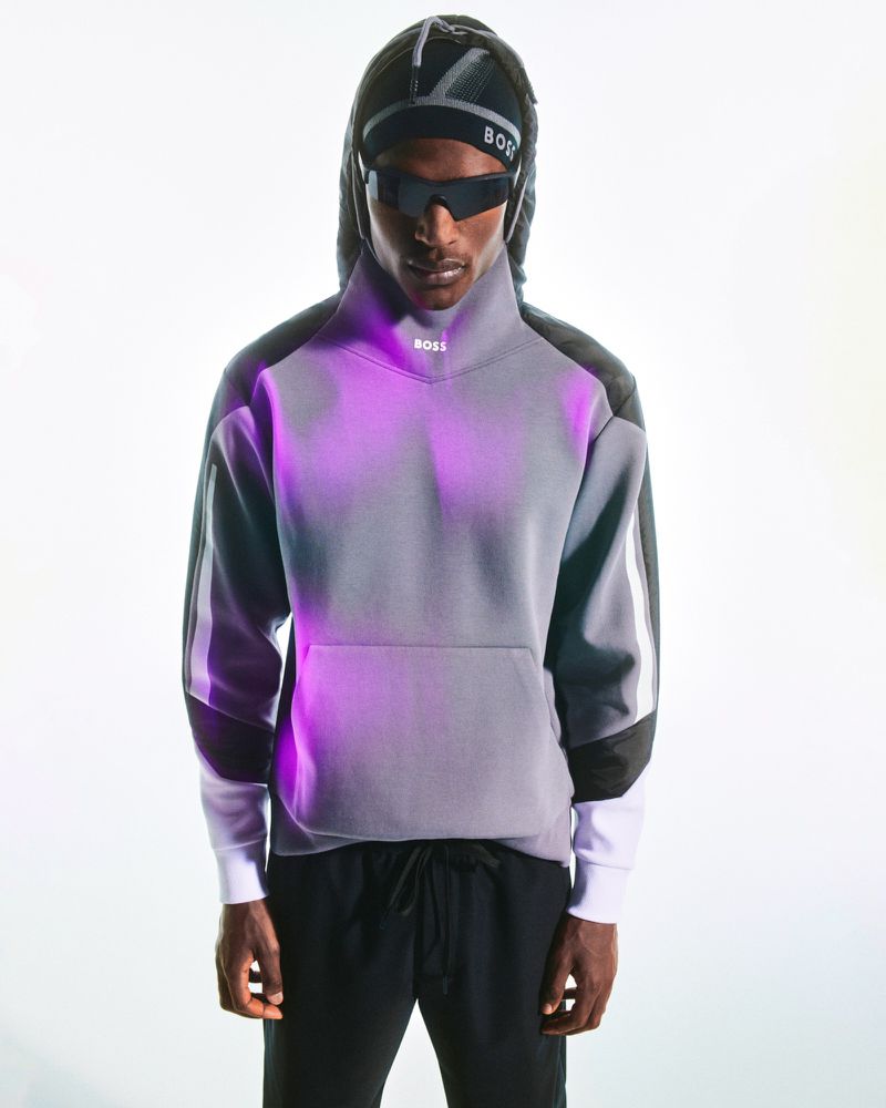 Portrait of a person wearing a grey sweatshirt, black sweatpants, sunglasses, and a hooded cap in a brightly lit indoor setting.