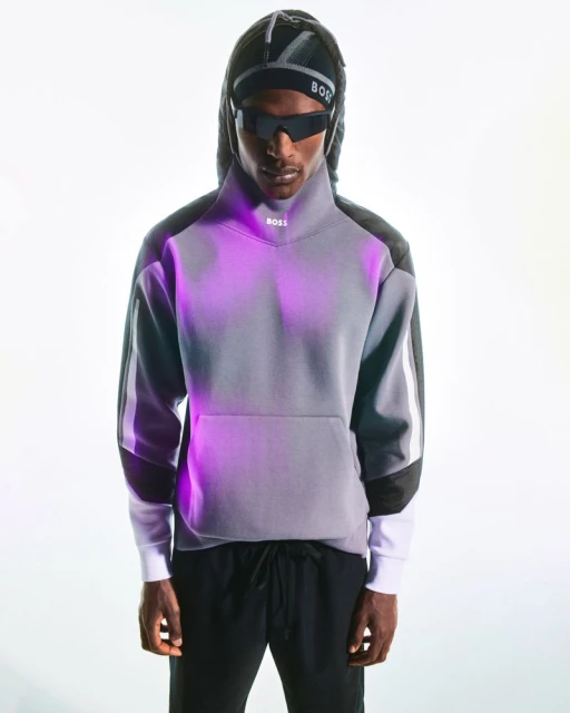 Portrait of a person wearing a grey sweatshirt, black sweatpants, sunglasses, and a hooded cap in a brightly lit indoor setting.
