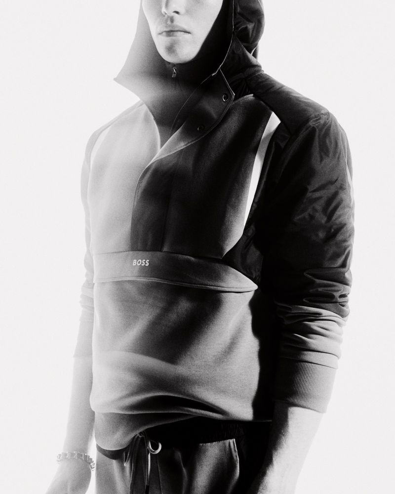 Monochrome portrait of a slender male model in a hooded sweatshirt and pants, featuring a subtle "BOSS" logo. Fashion-forward studio shot.
