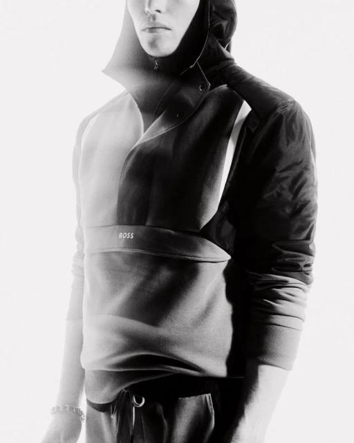Monochrome portrait of a slender male model in a hooded sweatshirt and pants, featuring a subtle "BOSS" logo. Fashion-forward studio shot.