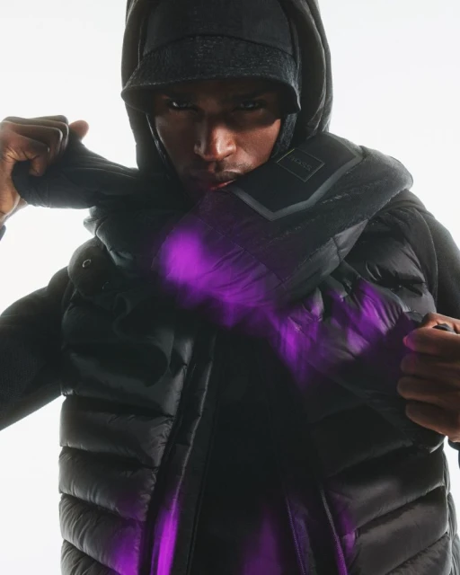 A person in a black hooded vest and cap poses with a serious expression against a bright white backdrop, featuring a futuristic purple light effect.