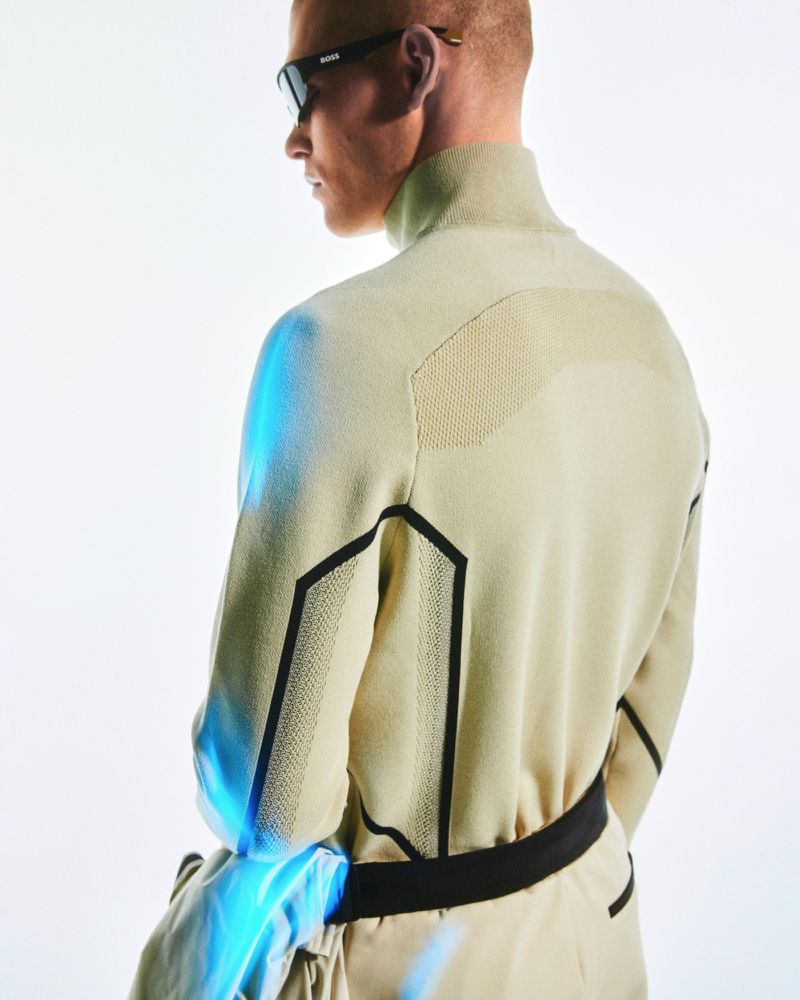 Man in beige high-neck sweater and matching pants wearing sunglasses in a bright studio environment. Fashion-forward sportswear.