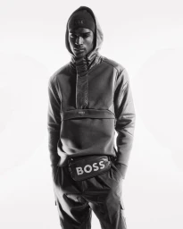 Model Rafael Mayers wearing a hooded technical pullover, beanie, and branded cross-body bag for BOSS Green in black and white.