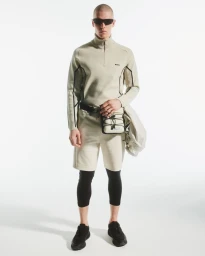 Model wearing a beige half-zip sweatshirt, matching shorts over black tights, sunglasses, and technical crossbody bag on white backdrop