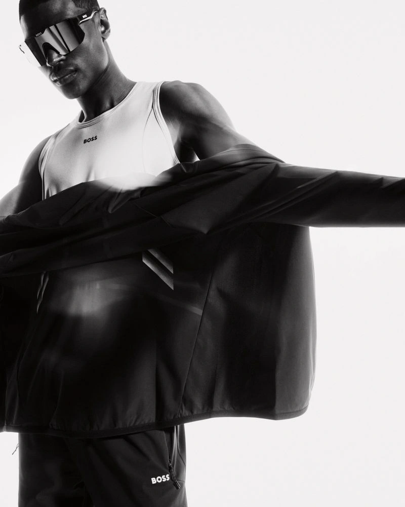 Model Rafael Mayers wearing athletic shield sunglasses, a white tank top, and a dark track jacket against a bright white background.