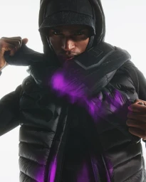 Man wearing a black hooded puffer jacket and baseball cap holding a padded wrap against a white seamless background