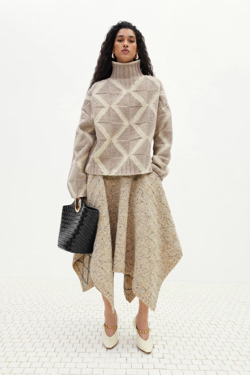 Elegant adult woman in beige patterned sweater and skirt holding black handbag, wearing heeled shoes, posing in studio lighting.