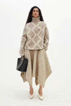 Elegant adult woman in beige patterned sweater and skirt holding black handbag, wearing heeled shoes, posing in studio lighting.