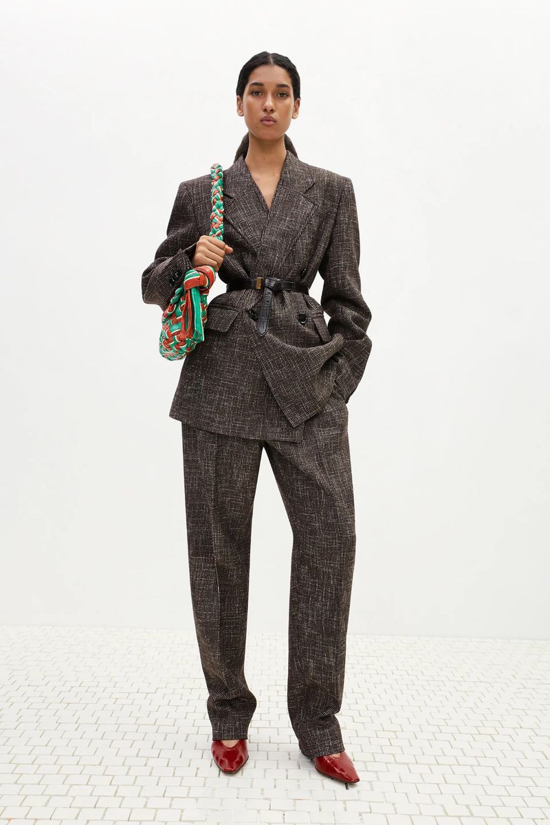Elegant woman in a tweed suit with shoulder bag and red shoes stands against a white background in a fashion studio shot.
