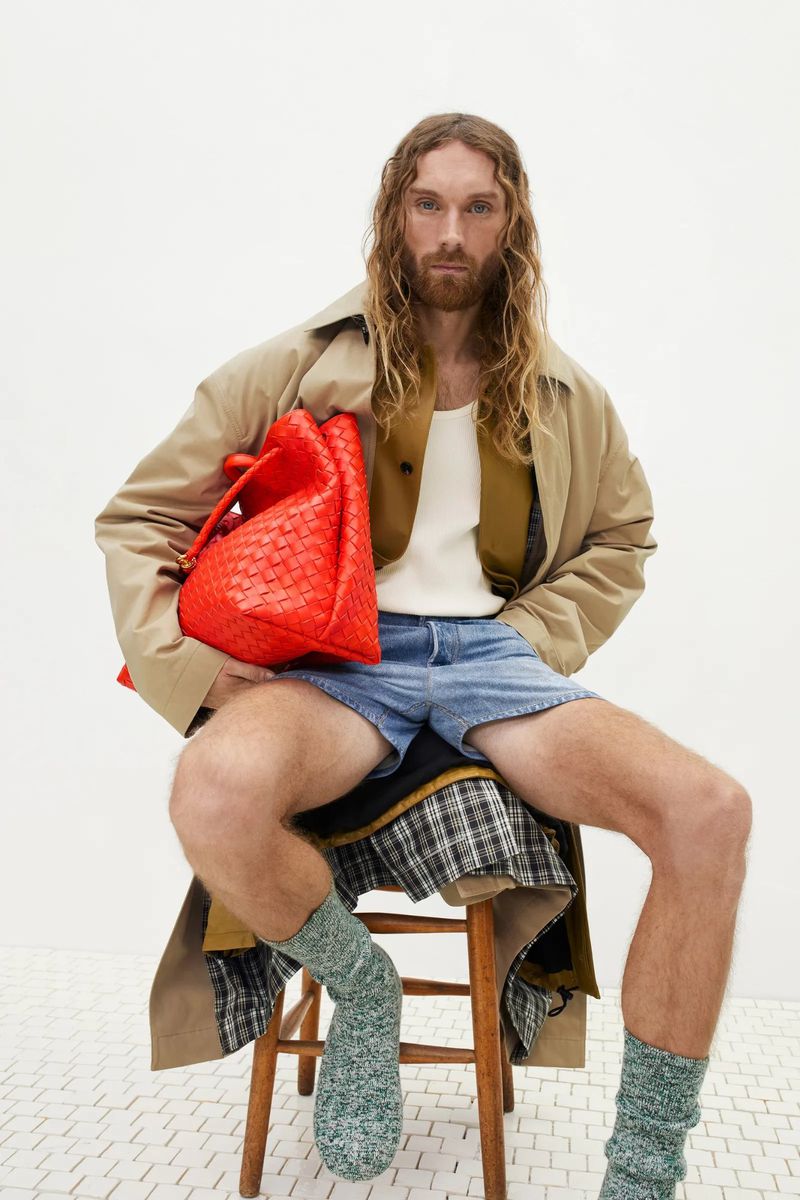 Man sits on a wooden stool in a studio with a red woven tote bag. He wears a trench coat, blue shorts, and green socks against a white backdrop.