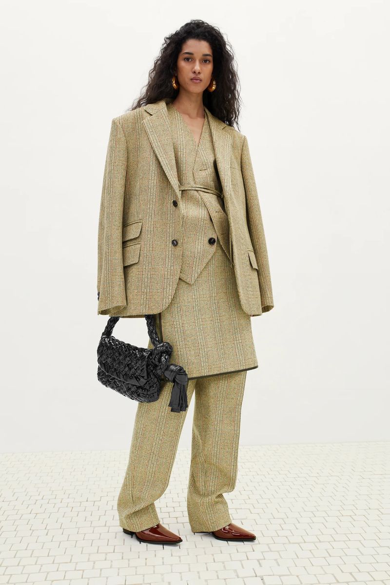 Adult female model wearing an oversized blazer and matching pants, holding a black purse, and posing in a photo studio.