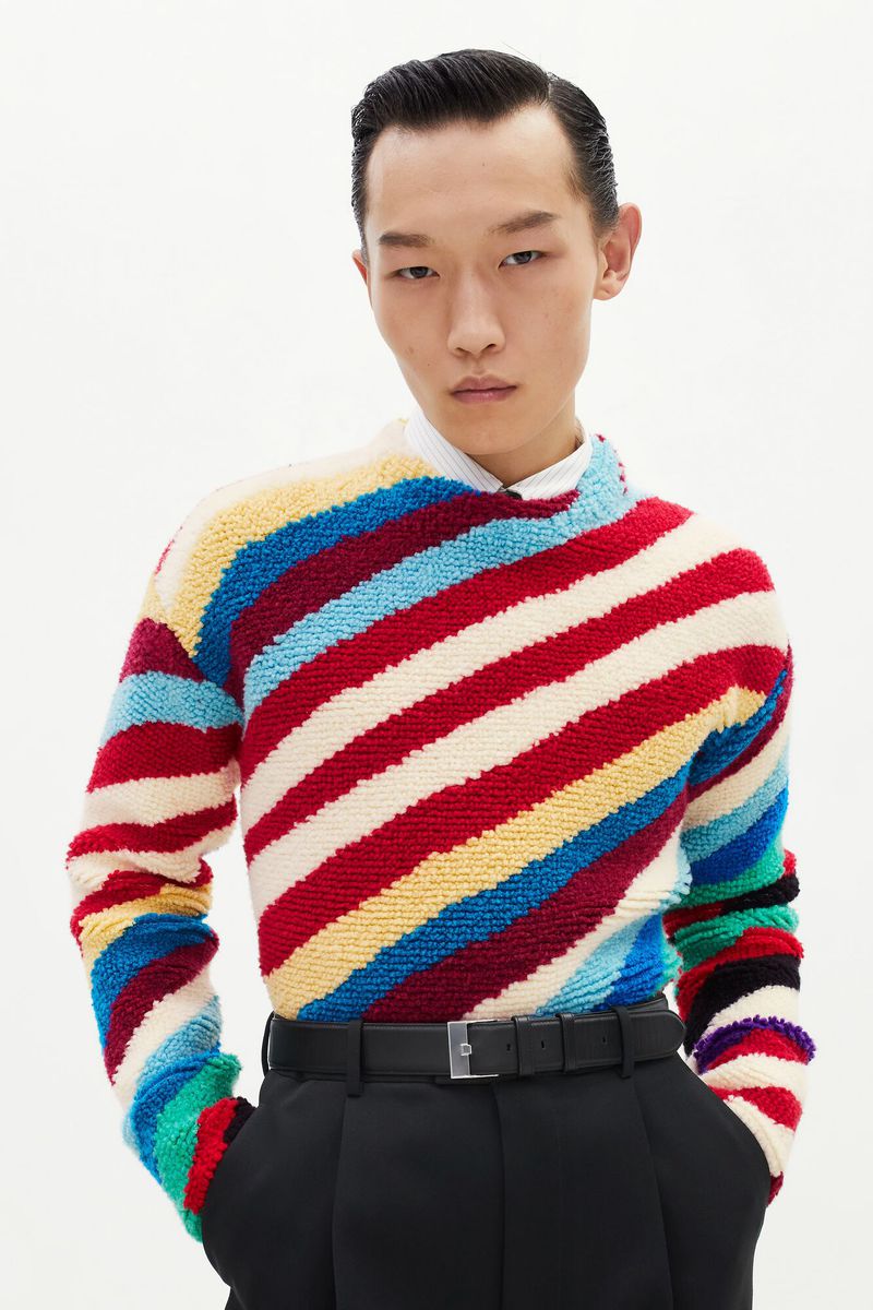 A young adult Asian man poses in a colorful striped knit sweater, black trousers, and belt against a bright, neutral backdrop.