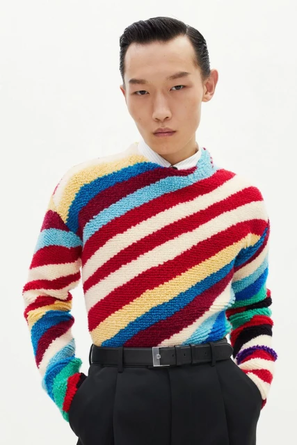 A young adult Asian man poses in a colorful striped knit sweater, black trousers, and belt against a bright, neutral backdrop.