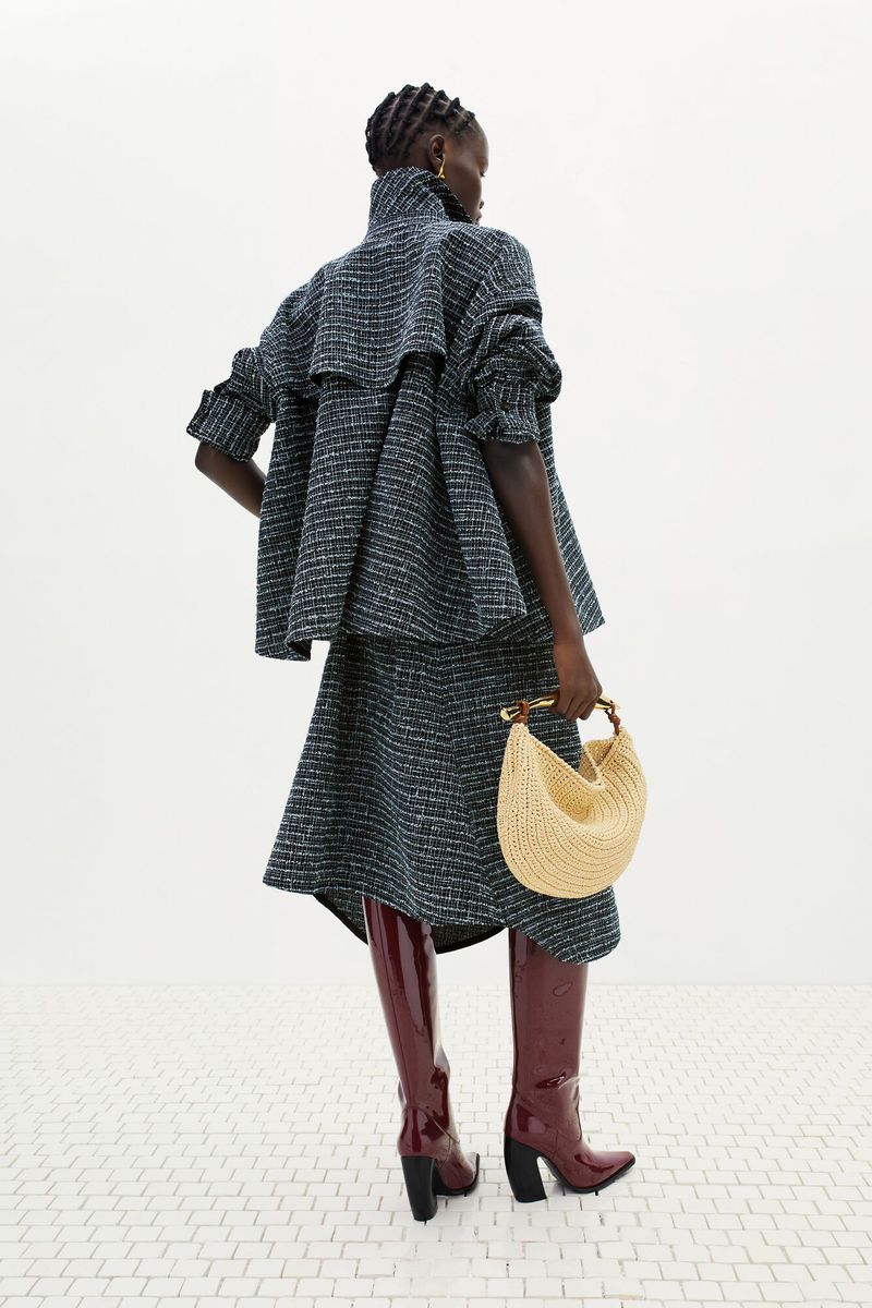 A woman wearing a tweed skirt suit and dark red patent leather boots holds a half-moon shaped straw purse with a light background.