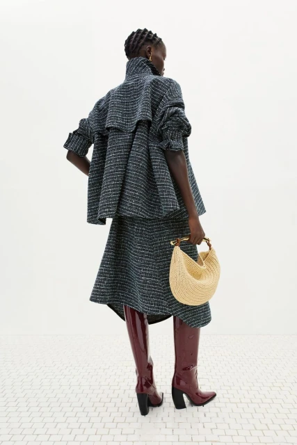 A woman wearing a tweed skirt suit and dark red patent leather boots holds a half-moon shaped straw purse with a light background.