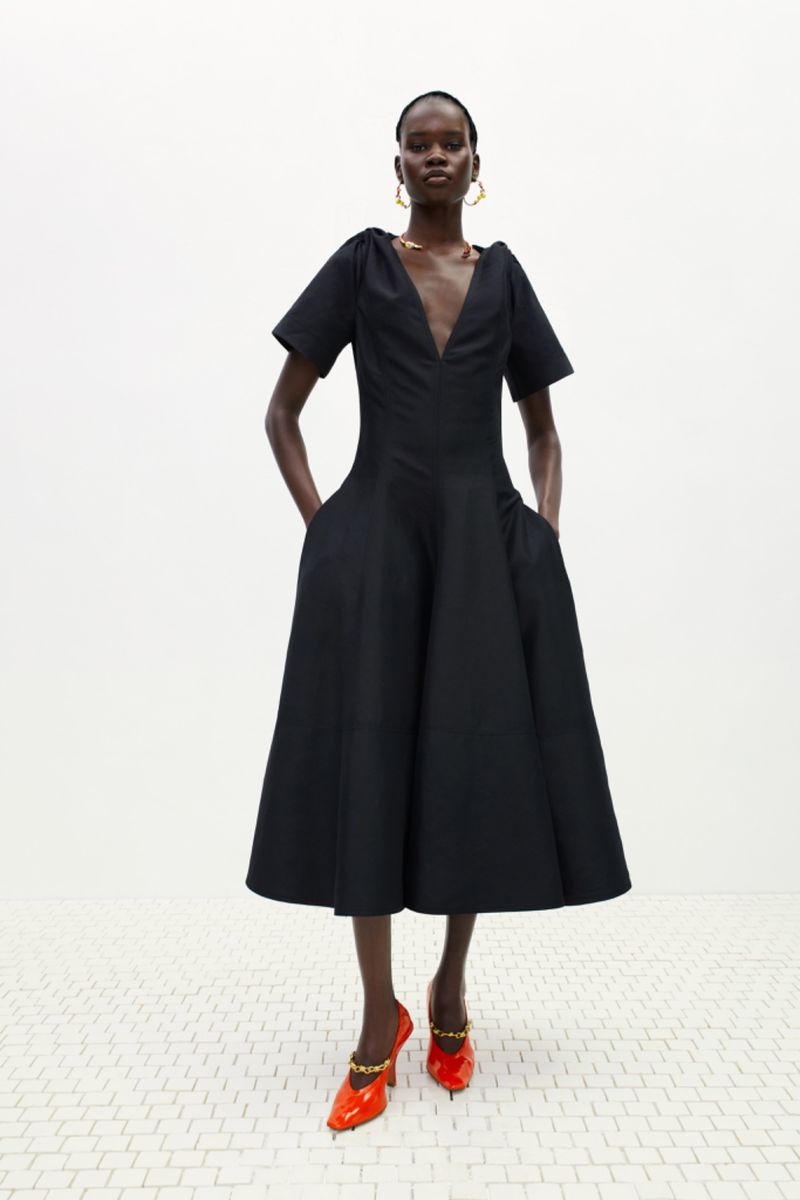 Full-length shot of a Black female fashion model wearing a black dress with orange high heels in a studio with a white background.