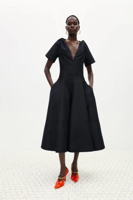 Full-length shot of a Black female fashion model wearing a black dress with orange high heels in a studio with a white background.