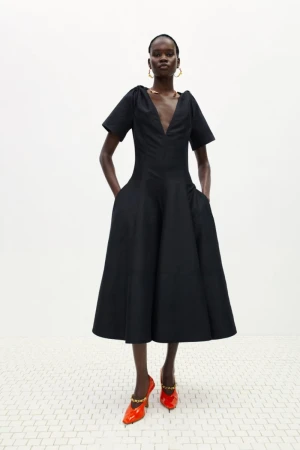 Full-length shot of a Black female fashion model wearing a black dress with orange high heels in a studio with a white background.