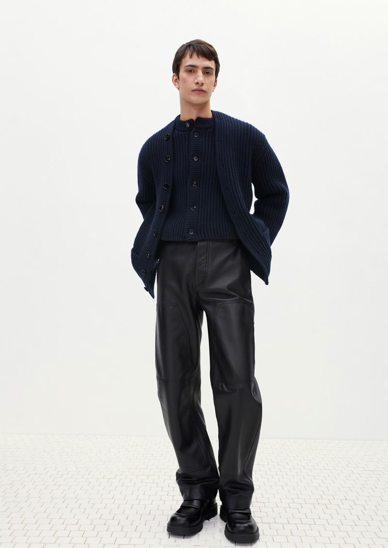 A stylish adult male poses in a fashion shot, wearing a navy knit cardigan, matching top, shiny black pants, and chunky black shoes against a plain white background.