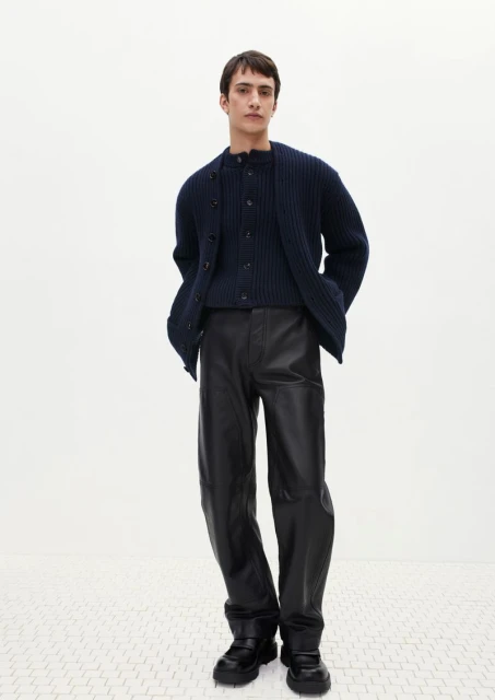 A stylish adult male poses in a fashion shot, wearing a navy knit cardigan, matching top, shiny black pants, and chunky black shoes against a plain white background.