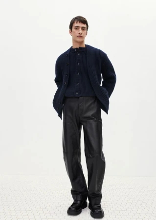 A stylish adult male poses in a fashion shot, wearing a navy knit cardigan, matching top, shiny black pants, and chunky black shoes against a plain white background.