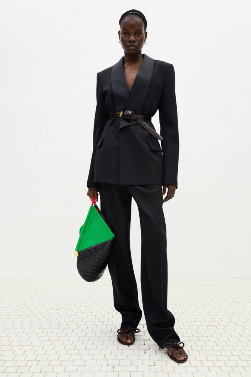 Model wearing a black belted tuxedo blazer with matching trousers and holding a green and black woven leather tote bag in a white studio.