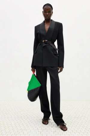 Model wearing a black belted tuxedo blazer with matching trousers and holding a green and black woven leather tote bag in a white studio.