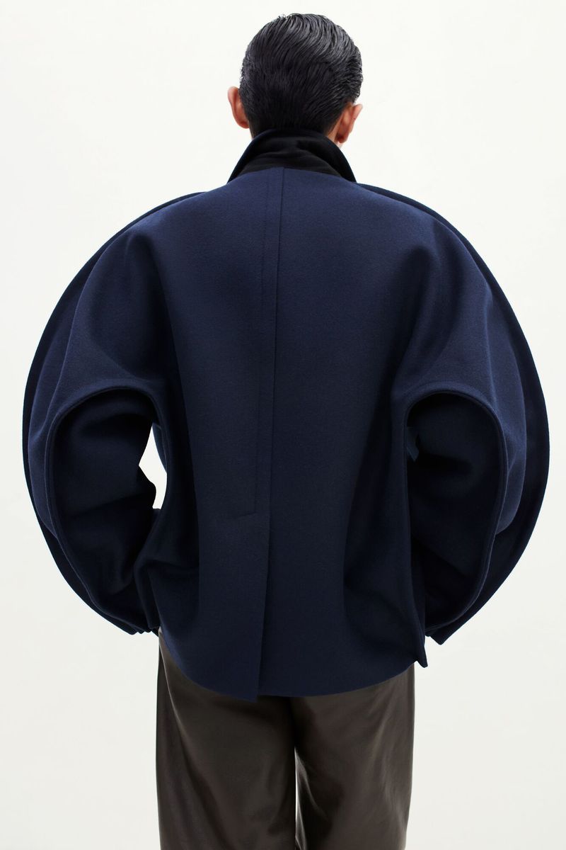 Rear view of a model wearing an exaggerated sculptural navy cocoon jacket with brown trousers against a white studio background.