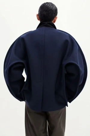 Rear view of a model wearing an exaggerated sculptural navy cocoon jacket with brown trousers against a white studio background.