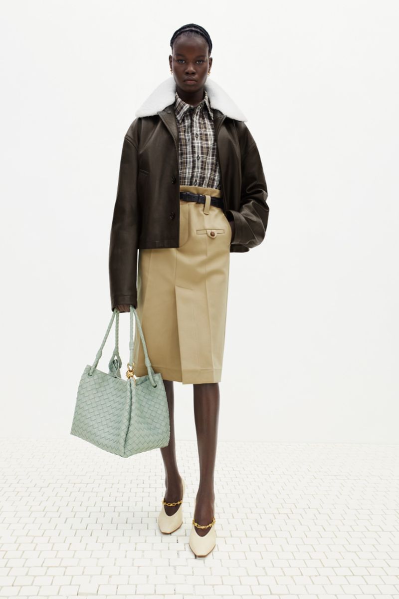 Fashion shoot of an African woman wearing a green jacket, plaid shirt, khaki skirt, and shoes holding a handbag against a white backdrop.