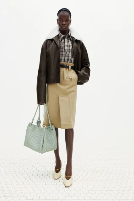 Fashion shoot of an African woman wearing a green jacket, plaid shirt, khaki skirt, and shoes holding a handbag against a white backdrop.