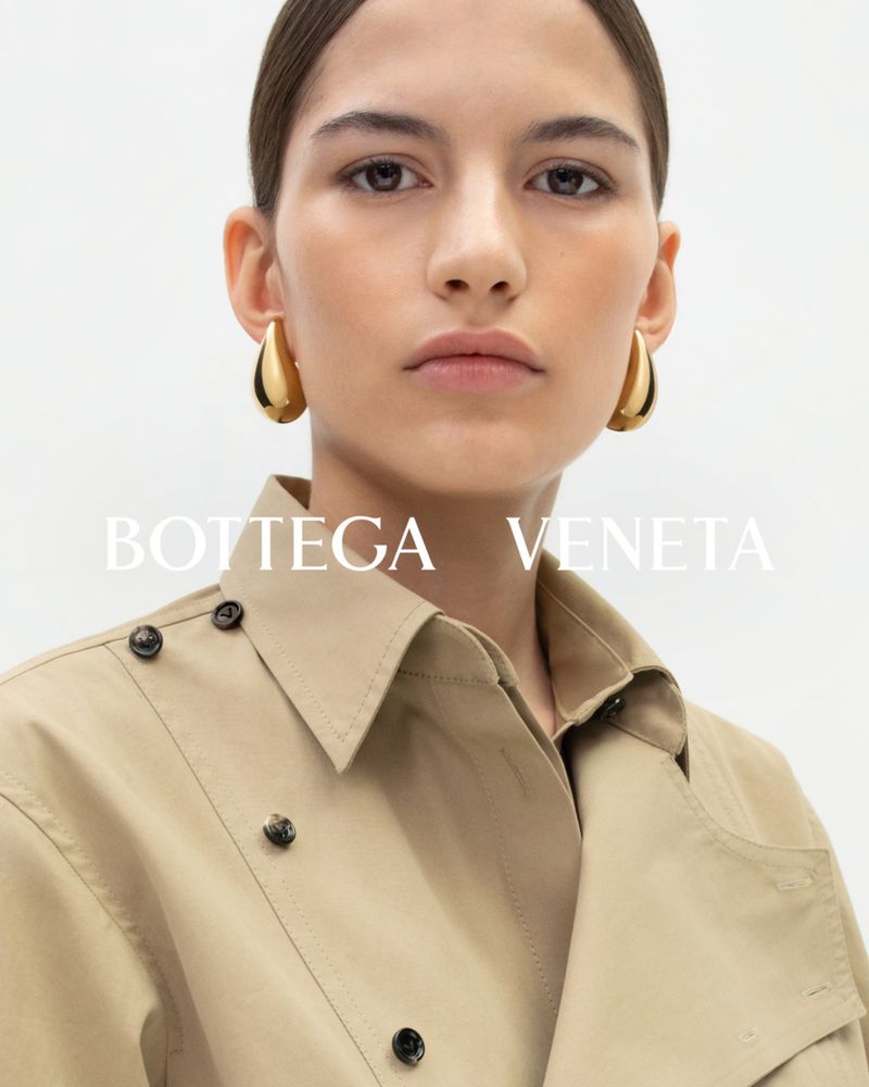 Close-up of a woman wearing gold hoop earrings and a tan collared coat in a studio setting, showcasing a fashion editorial look.