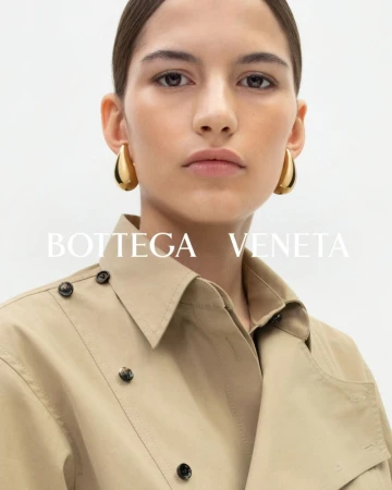 Close-up of a woman wearing gold hoop earrings and a tan collared coat in a studio setting, showcasing a fashion editorial look.