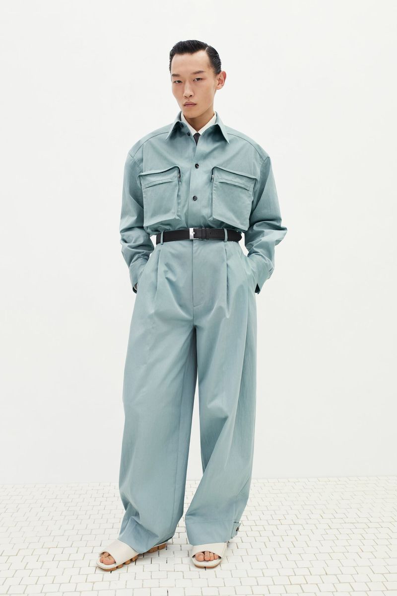 A full-length shot of an adult male fashion model in a light blue two-piece outfit, black belt, and light-colored sandals against a white backdrop.
