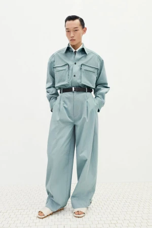 A full-length shot of an adult male fashion model in a light blue two-piece outfit, black belt, and light-colored sandals against a white backdrop.