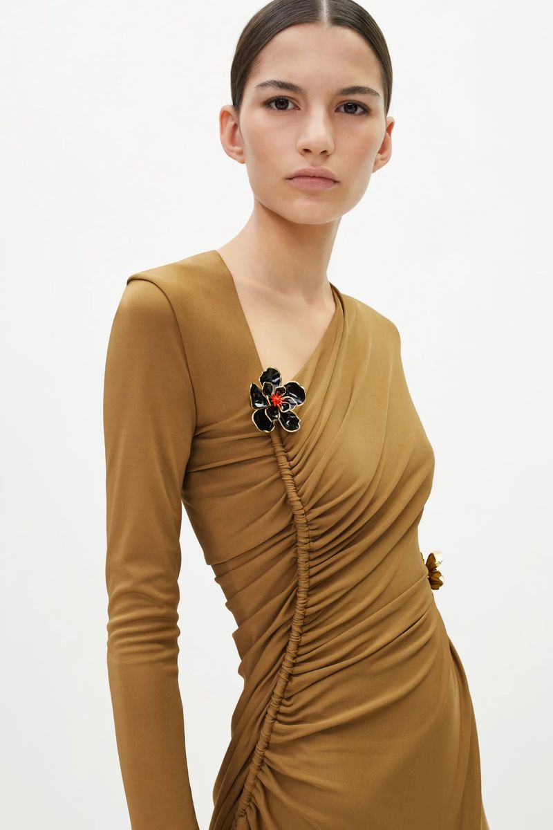 A woman wearing a long-sleeved dress with flower brooches poses against a white backdrop in a studio setting with soft lighting.