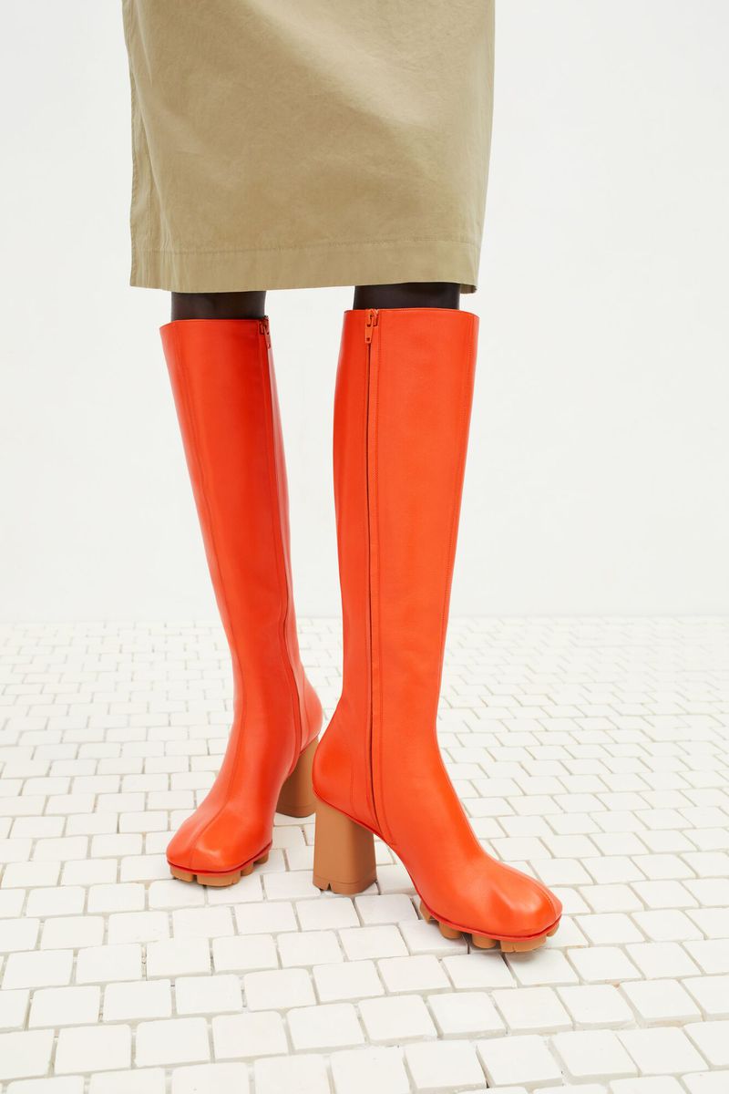 Close-up of orange knee-high boots with a tan block heel on a white brick surface, complemented by a tan skirt or garment.