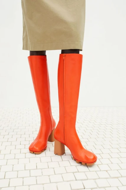 Close-up of orange knee-high boots with a tan block heel on a white brick surface, complemented by a tan skirt or garment.