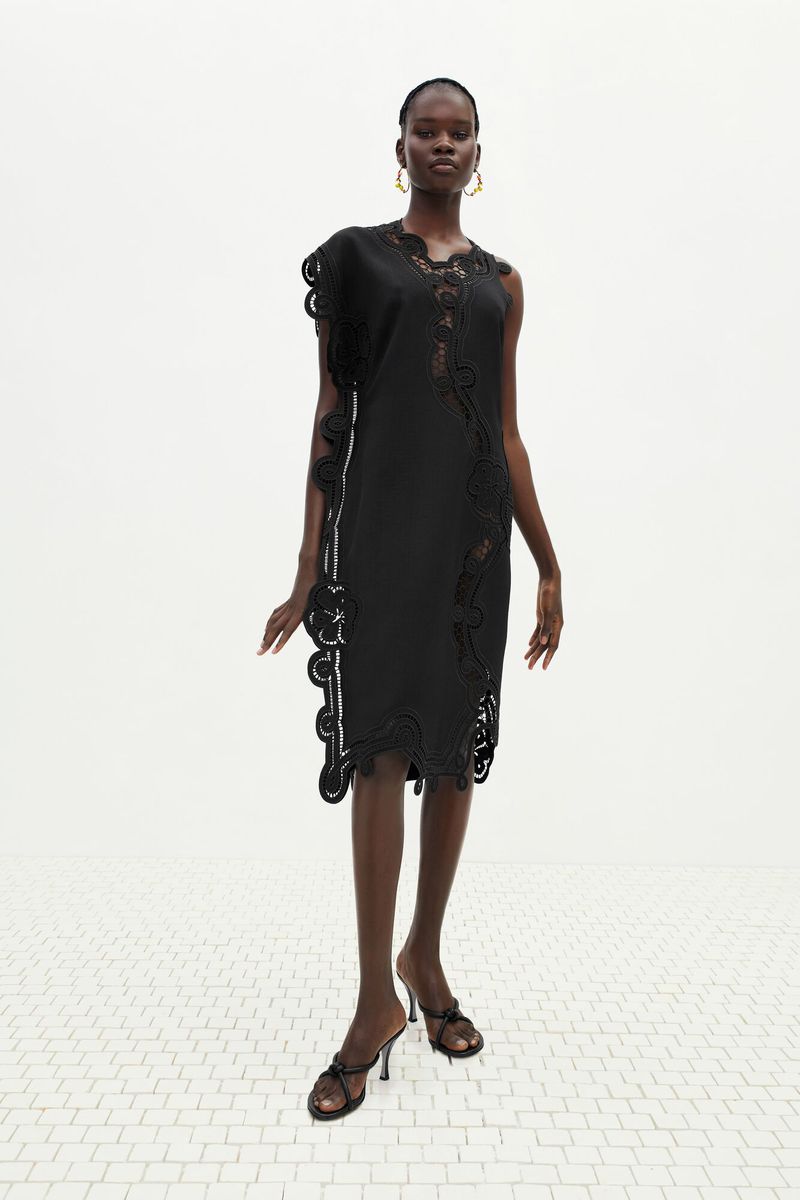 Fashion shoot featuring a slender, dark-skinned woman wearing a black dress with decorative trim and black high-heeled shoes against white backdrop.