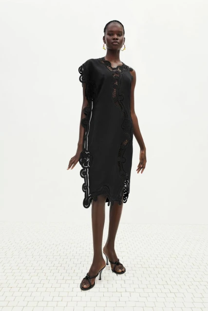 Fashion shoot featuring a slender, dark-skinned woman wearing a black dress with decorative trim and black high-heeled shoes against white backdrop.