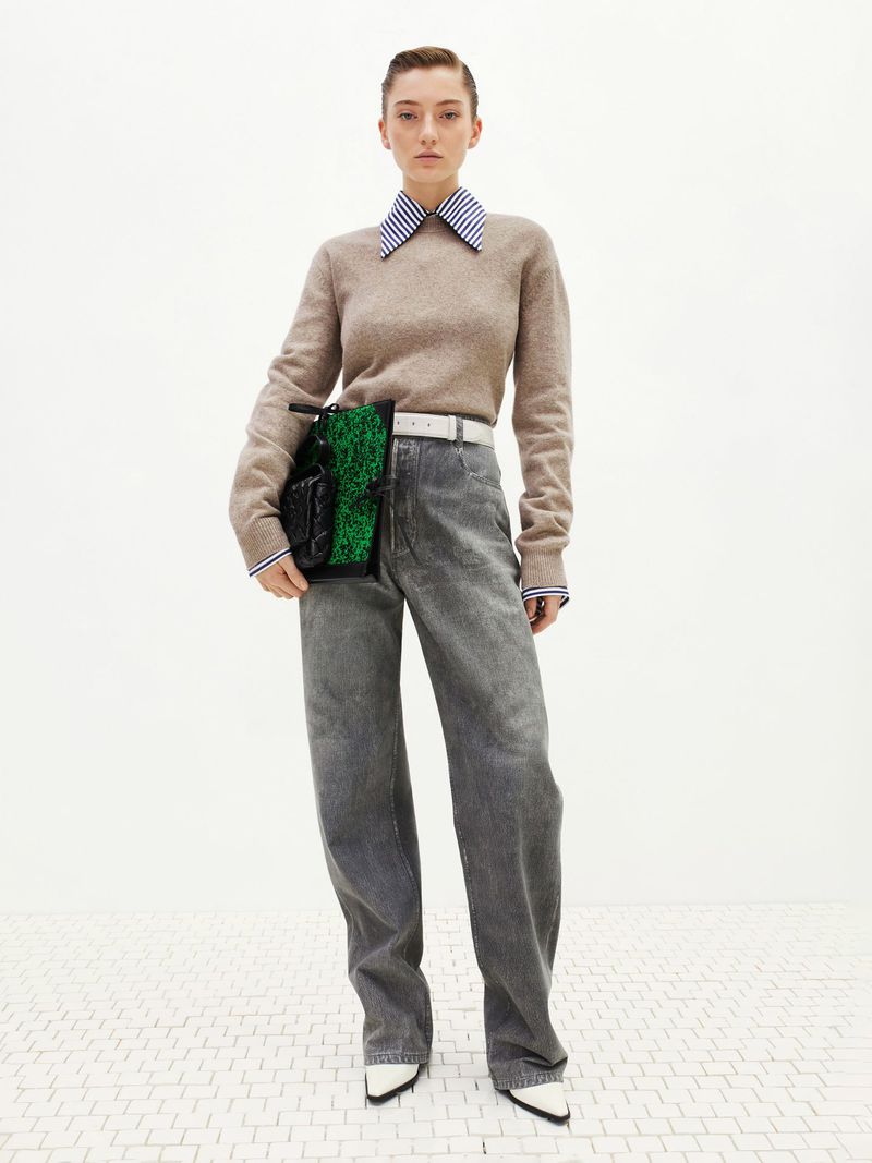 A young adult woman poses in a studio wearing wide-leg denim jeans, a beige sweater, and a green and black handbag.