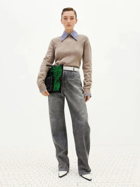 A young adult woman poses in a studio wearing wide-leg denim jeans, a beige sweater, and a green and black handbag.