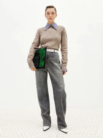 A young adult woman poses in a studio wearing wide-leg denim jeans, a beige sweater, and a green and black handbag.