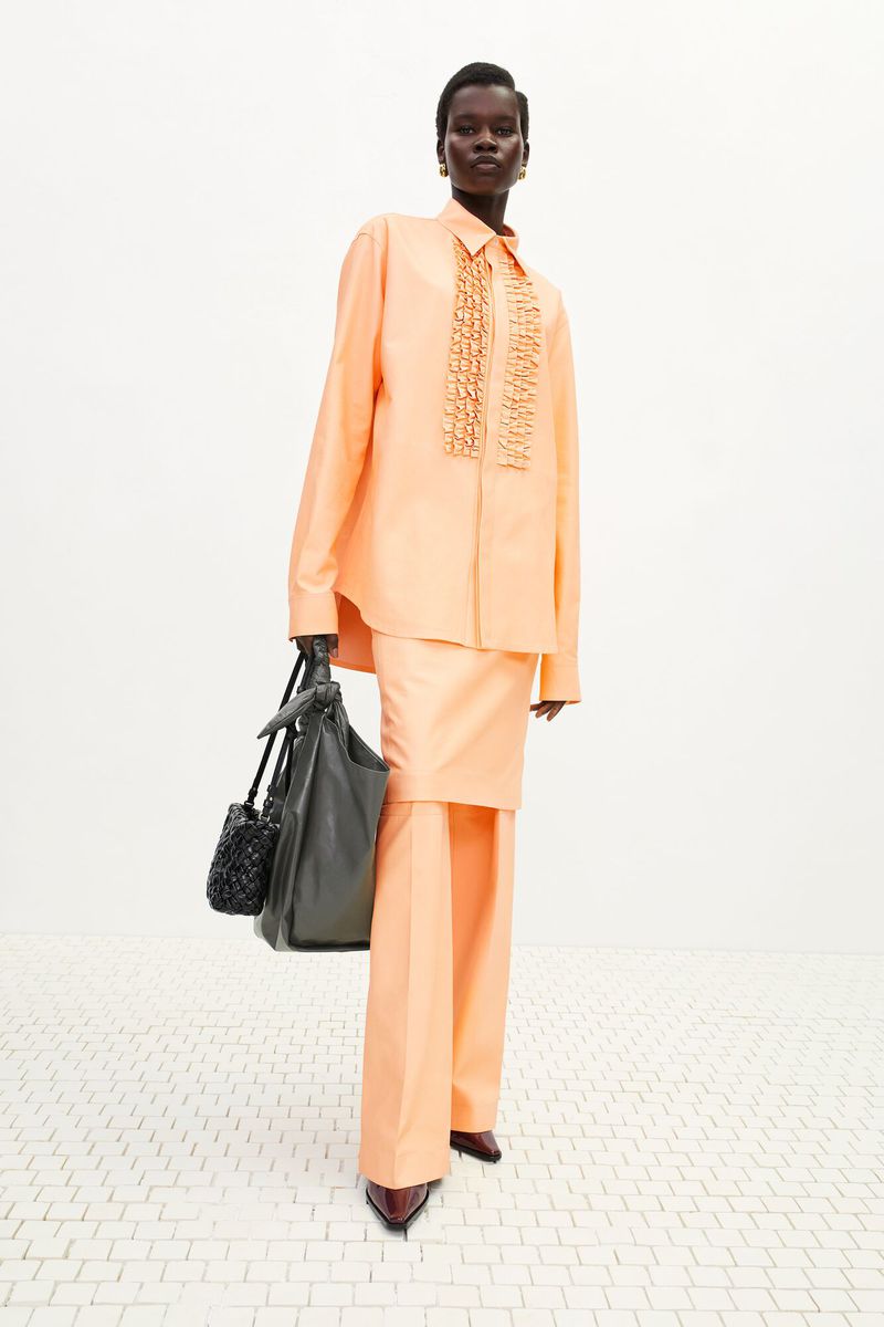 Woman in peach suit with matching accessories, standing in a studio with simple white flooring and backdrop, full-length, eye-level angle.