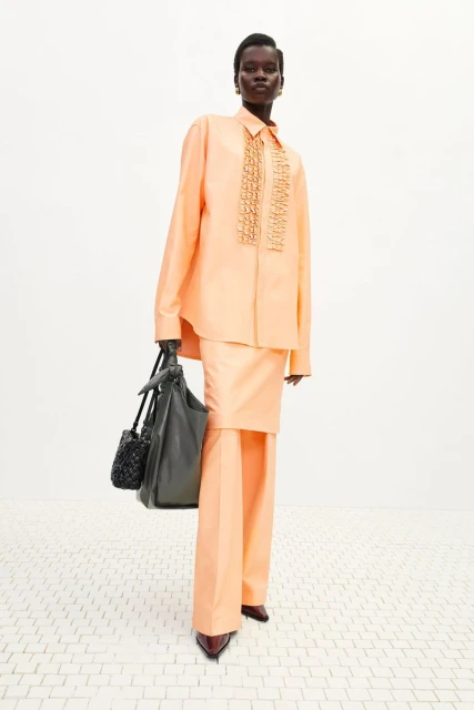 Woman in peach suit with matching accessories, standing in a studio with simple white flooring and backdrop, full-length, eye-level angle.