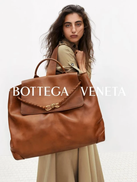Fashion portrait of a brunette woman holding a large brown leather Bottega Veneta handbag with a white studio background.