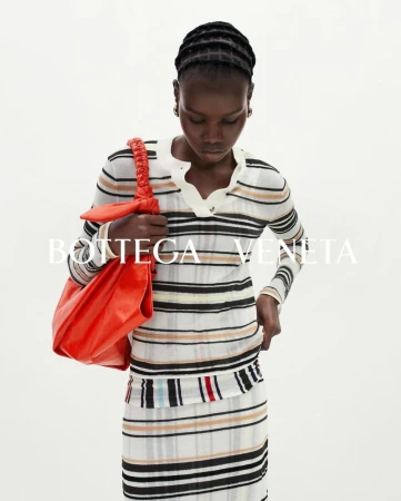 Model wearing a striped long-sleeve knit top and matching skirt while carrying a vibrant orange leather shoulder bag against a white backdrop.