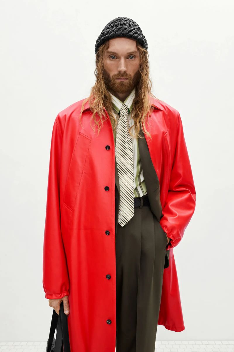 Portrait of a long-haired male model in a red leather coat, striped shirt, and tie against a plain white background in a studio setting.