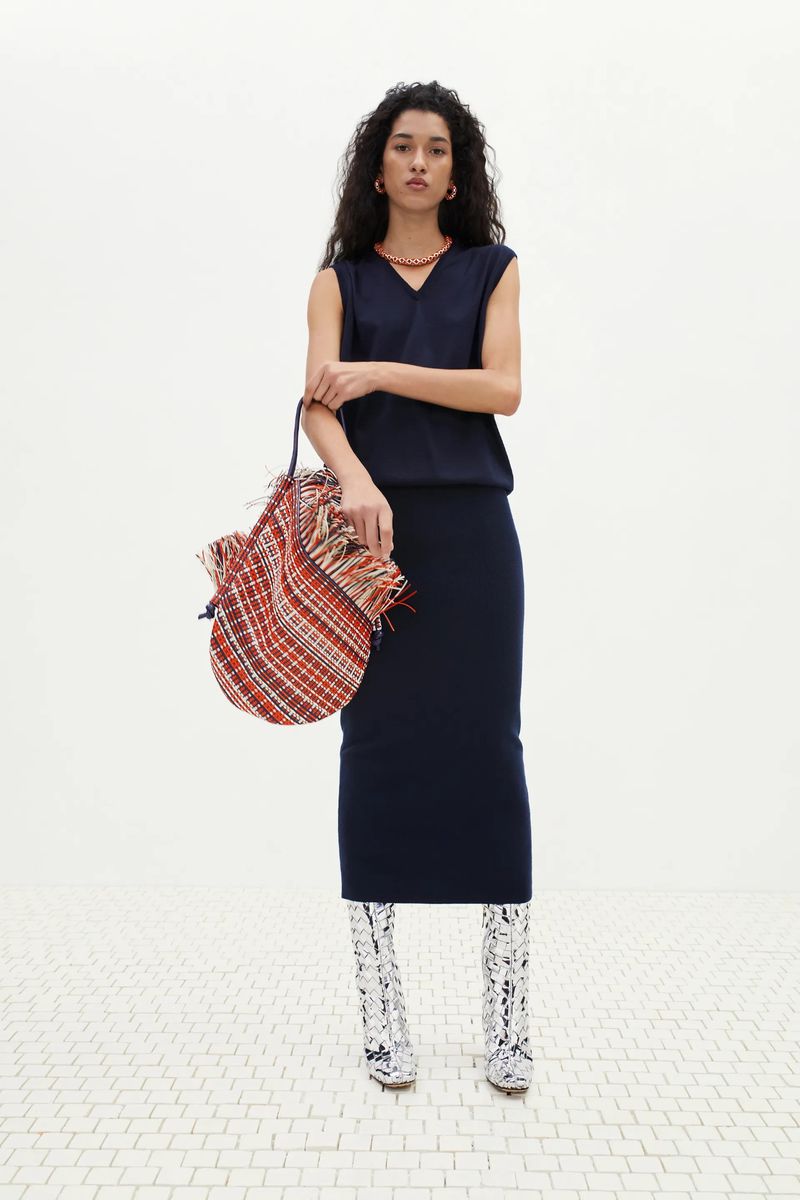 Full shot of a woman posing in a sleeveless dark blue top and pencil skirt, holding a colorful, patterned bag, set against a white background.