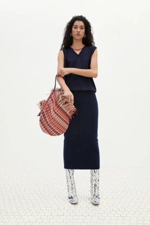 Full shot of a woman posing in a sleeveless dark blue top and pencil skirt, holding a colorful, patterned bag, set against a white background.